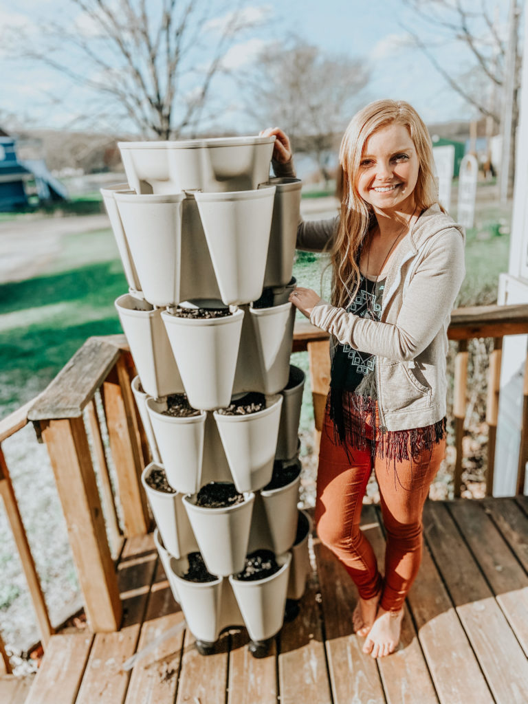 blonde girl (kiersten Zile) standing next to a greenstalk vertical garden tower smiling at the camera standing on a porch with a lake behind the her and a barn