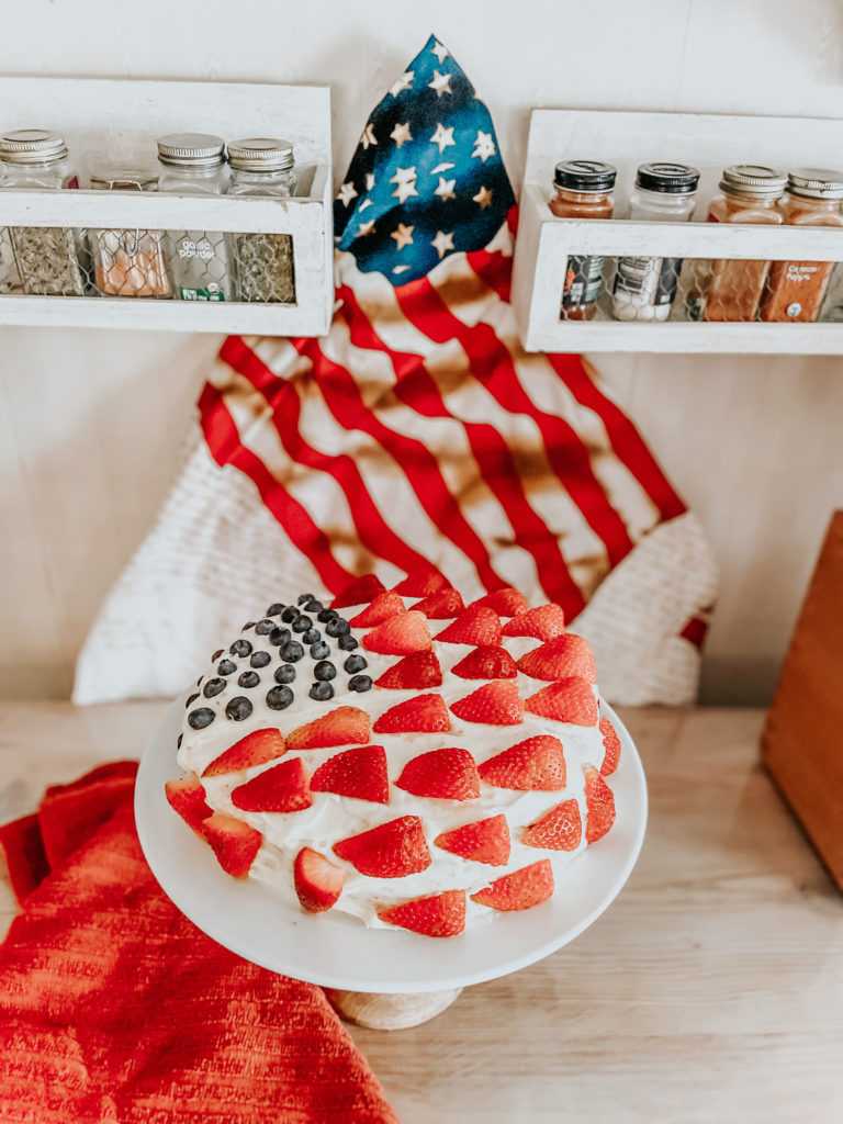 a double layered vanilla einkorn cake with strawberries and blueberries in the shape of an American flag with an American flag behind the cake