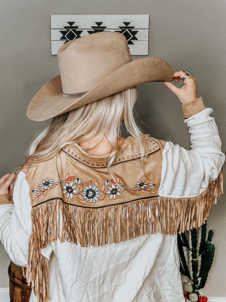 kiersten zile holding her hair up with a fringe western button-up from a back view to show the fringe and flower detail on the western fall dress style with a tan cowboy hat on
