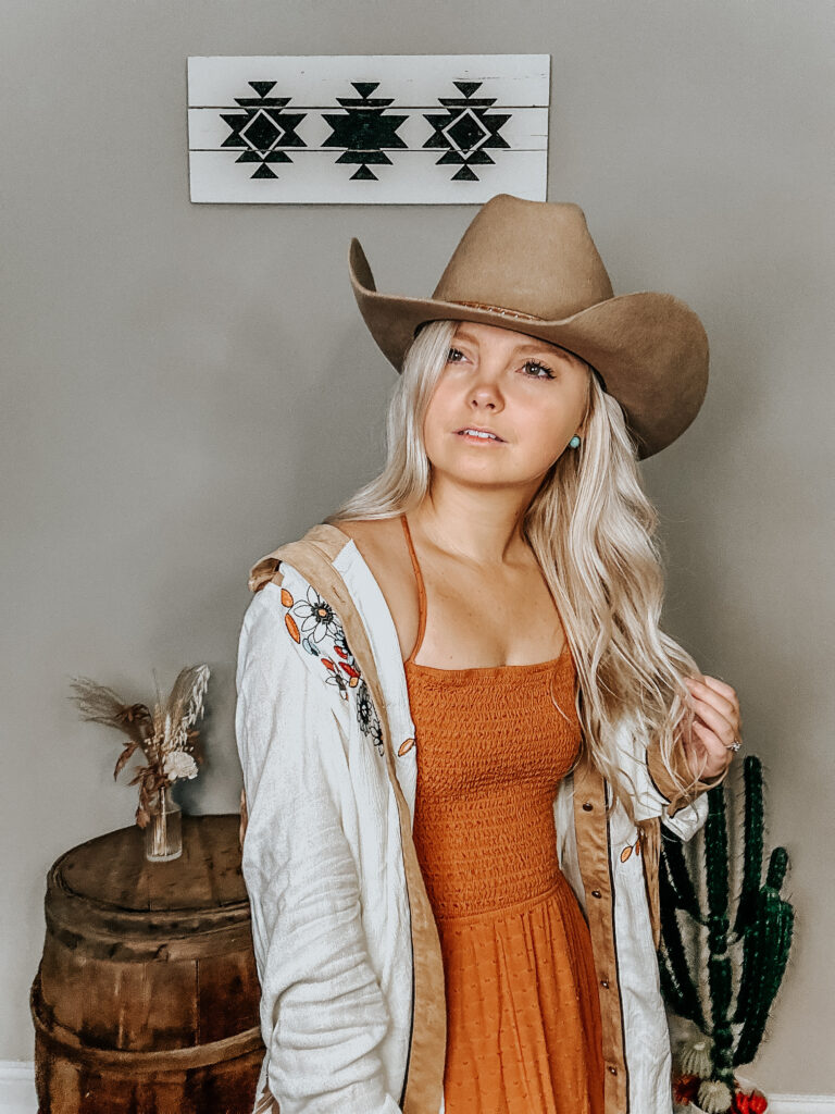 kiersten zile holding her hair up with a fringe western button-up and a burnt orange dress to show the western fall dress style with turquoise earrings on and a felt western cowboy hat